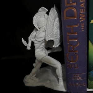 May include: A white figurine of a warrior in a helmet and shield, holding a knife, in a dynamic pose. The figurine is positioned next to a book with the title "EARTH DRAGON". The warrior is on a small base.