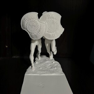 May include: A white 3D-printed sculpture of two figures holding shields. The shields have a sunburst design. The figures are on a textured base, with a rectangular platform below.