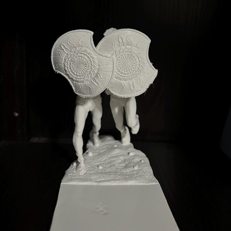 May include: A white 3D-printed sculpture of two figures holding shields. The shields have a sunburst design. The figures are on a textured base, with a rectangular platform below.