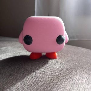 May include: A pink 3D printed figurine of a character with large black eyes and red feet. The figurine is in a sitting position with its legs spread out.