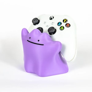 May include: A purple Ditto-shaped controller holder supporting a white Xbox controller. The controller has black joysticks and buttons with colourful A, B, X, and Y buttons. The Ditto figure has a simple black smile and dot eyes.