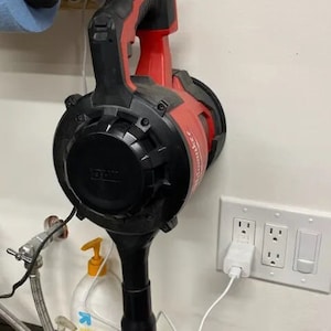 May include: A red and black cordless leaf blower plugged into an electrical socket. The blower is mounted on a wall with a white cord.