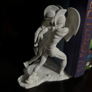 May include: A white sculpture of two ancient Greek warriors in battle. One warrior holds a shield and the other appears to be lunging forward. The sculpture is detailed and stands on a small base, perfect for a bookshelf.