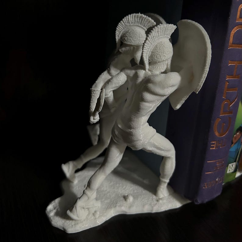 May include: A white sculpture of two ancient Greek warriors in battle. One warrior holds a shield and the other appears to be lunging forward. The sculpture is detailed and stands on a small base, perfect for a bookshelf.