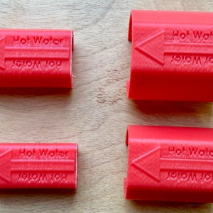 May include: Four red plastic clips, each with the words "Hot Water" and an arrow indicating direction. The text is embossed on the surface of the clips. The clips are arranged on a light brown wooden surface.