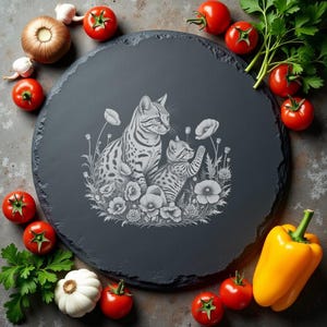 Puede incluir: Una tabla redonda de pizarra gris oscuro con un diseño grabado con láser de dos gatos manchados entre flores. La tabla está rodeada de tomates frescos, ajo, un pimiento amarillo y perejil.