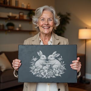 以下が含まれることがあります： 暗い灰色の長方形のスレートボード。3匹のウサギが花に囲まれた詳細な彫刻が施されています。ウサギは家族として描かれ、2匹の大きなウサギと1匹の小さなウサギがいます。アートワークはグレースケールです。