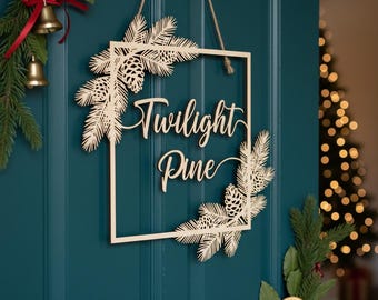 May include: A wooden sign with the words "Twilight Pine" in a decorative font. The sign is square with pine branches and pinecones in each corner. It hangs from a natural twine string. The sign is displayed on a teal door.
