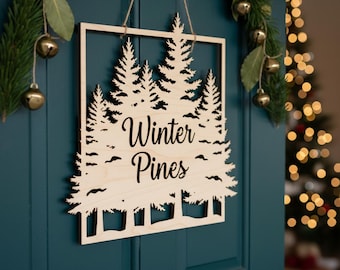 May include: A wooden sign with the words "Winter Pines" cut out in a decorative font. The sign features a forest silhouette and is framed by a square border. The sign is hanging on a teal door, with Christmas decorations in the background.