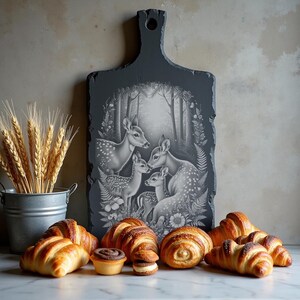 Op de afbeelding: Een donkergrijze snijplank met een handvat, met een zwart-wit boslandschap met herten. Vers gebakken croissants en gebakjes zijn voor het bord gerangschikt, met een kleine metalen emmer met tarwestengels aan de linkerkant.