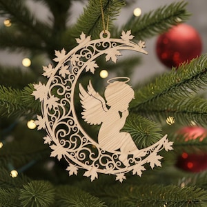 May include: Wooden Christmas ornament featuring an angel seated on a crescent moon. The ornament is made of light-colored wood and has intricate cut-out details, including leaves and swirling patterns. A gold string is attached for hanging.