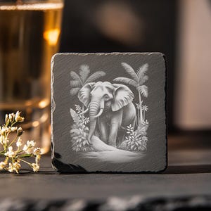 May include: Square slate coaster featuring an engraved image of an elephant in a jungle setting. The elephant is surrounded by palm trees and lush foliage. The coaster is dark gray and has a natural, textured surface.