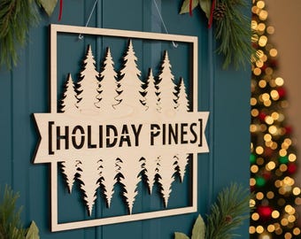 May include: A wooden sign with the text "[HOLIDAY PINES]" and a cutout of pine trees. The sign is rectangular with a square frame and is hanging on a teal door. Christmas decorations, including bells and greenery, are visible.