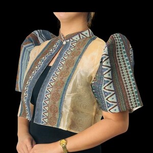 Può includere: Giacca bolero beige con maniche e finiture fantasia. La giacca presenta un collo alto e un design decorativo nei toni del blu, marrone e beige. Le maniche sono corte e a sbuffo. La modella indossa un top e una gonna neri.