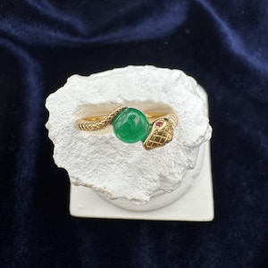 Pode incluir: Um anel de cobra de ouro com uma gema verde e um olho de rubi vermelho. O anel está em um suporte branco.