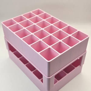 Può includere: Due vassoi portaoggetti rosa chiaro impilati, con fori quadrati. Ogni vassoio presenta una griglia di scomparti quadrati. I vassoi sono realizzati in un materiale solido e opaco, progettati per organizzare piccoli oggetti.