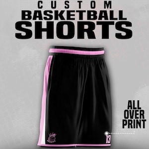 May include: Black basketball shorts with pink and white accents. The shorts have a pink waistband and side stripes. The text "CUSTOM BASKETBALL SHORTS" is at the top, and "ALL OVER PRINT" is on the side. The number "13" is on the shorts.