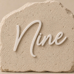 May include: A beige stone with the word "Nine" in white script. The stone has a rough, textured surface and a rectangular shape. The white lettering is in a cursive font.