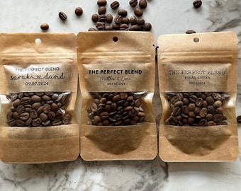 May include: Three brown paper bags with clear windows, each containing coffee beans and a label that reads "The Perfect Blend" with a wedding date and the couple's names. The bags are arranged in a row on a white marble surface.