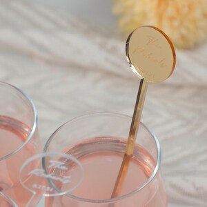 Puede incluir: Un agitador de bebidas de acr&iacute;lico dorado con una parte superior circular que presenta texto grabado en cursiva, colocado dentro de un vaso transparente lleno de un l&iacute;quido rosa claro. Se ve otro agitador transparente con texto blanco.