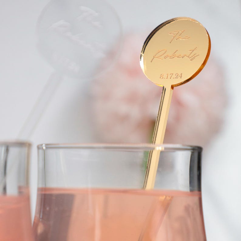 Puede incluir: Un agitador de bebidas de acr&iacute;lico dorado con una parte superior circular que presenta el texto grabado The Roberts y la fecha 8.17.24. El agitador est&aacute; colocado dentro de un vaso con l&iacute;quido rosa claro, con otro agitador transparente borroso detr&aacute;s.