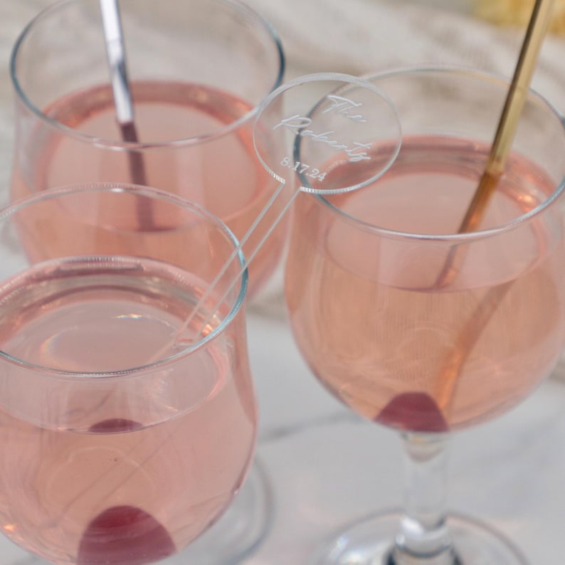Puede incluir: Tres copas de cristal transparente llenas de un l&iacute;quido rosa claro con una cereza roja oscura en el fondo de cada una. Se incluye un agitador de bebidas circular de acr&iacute;lico transparente con el texto grabado The Roberts 8.17.24.
