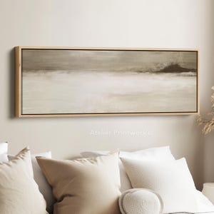 Op de afbeelding: Abstract landschapsschilderij in neutrale tinten, ingelijst in licht hout. Het kunstwerk heeft een horizontale compositie met een mix van beige, bruin en wit. De tekst "Atelier Printworks." is zichtbaar onderaan.