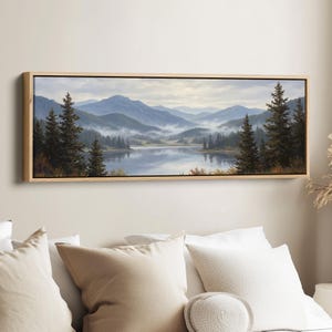May include: A landscape painting in a light wood frame depicts a serene mountain lake scene. The artwork features misty blue mountains, evergreen trees, and the reflection of the landscape in the calm water. The painting is rectangular and horizontal.