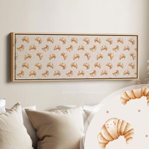 Peut inclure: Impression sur toile rectangulaire avec un motif répété de croissants à l'aquarelle sur fond crème. Les croissants sont dans les tons bruns et beiges, avec de petits points bruns. Encadrement en bois clair.
