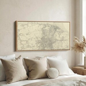 May include: A framed vintage map of Edinburgh, Scotland, in shades of gray and beige. The map is rectangular and mounted in a light-colored wooden frame. The map is displayed above a bed with beige and white pillows.