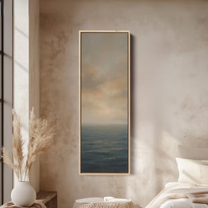 Op de afbeelding: Een rechthoekig ingelijst kunstwerk toont een zeegezicht met een gedempt kleurenpalet. Het schilderij toont een donkerblauwe oceaan die een mistige, lichtgekleurde lucht ontmoet. Het kunstwerk is ingesloten in een lichtgekleurde houten lijst, wat een serene en minimalistische esthetiek creëert.