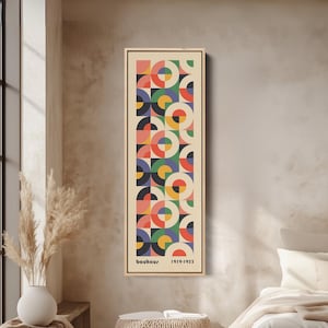 May include: A vertical rectangular wall art print in a light wood frame featuring a repeating geometric pattern of circles and semicircles in red, blue, green, yellow, and black on a cream background with the text Bauhaus 1919-1933.