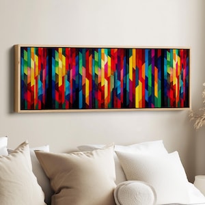 May include: A framed artwork with a wooden surround. The abstract design features vertical, colourful geometric shapes in red, yellow, blue, and green. The artwork is displayed above white pillows on a bed.