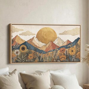 Op de afbeelding: Een ingelijste kunstdruk met een berglandschap, een grote gouden zon en een veld met wilde bloemen. Het kleurenpalet omvat tinten geel, oranje, blauw en beige. Het kunstwerk is in een houten lijst.