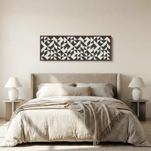 Puede incluir: Una pieza de arte mural rectangular con un patr&oacute;n geom&eacute;trico repetitivo de tri&aacute;ngulos y cuadrados en negro y blanco roto, encerrada en un marco de madera fino de color marr&oacute;n oscuro, montada horizontalmente sobre una pared beige claro.