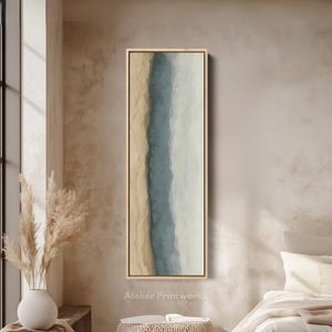 May include: A framed abstract art print with a vertical design. It features a central band of blue-grey, bordered by beige and off-white tones, within a light wood frame. The artwork suggests a coastal or landscape theme. The text "Atelier Printworks" is visible.