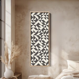 Könnte beinhalten: Ein hohes, rechteckiges Wandbild mit einem sich wiederholenden geometrischen Muster aus dunkel anthrazitfarbenen und cremeweißen Dreiecken und Quadraten, eingefasst in einen dünnen, hellen Holzrahmen.