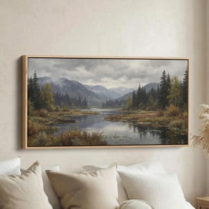 May include: A framed landscape painting showing a tranquil lake scene with mountains in the background. The artwork has a wooden frame and depicts a calm lake surrounded by trees and foliage, with a cloudy sky above.
