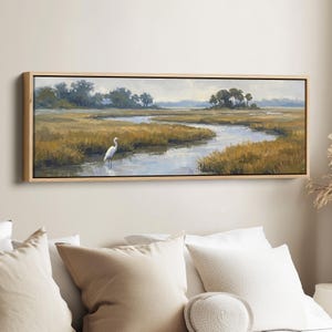 May include: A framed landscape painting depicts a serene marsh scene. The artwork features a winding river, golden grasses, and a white heron. The sky is overcast, and the painting is encased in a light-colored wooden frame.