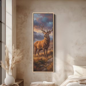 Puede incluir: Pintura vertical enmarcada de un majestuoso ciervo con grandes cuernos, de pie en un paisaje montañoso. El ciervo tiene pelaje marrón y se encuentra sobre un fondo de una colorida puesta de sol con tonos de naranja, morado y azul. La obra de arte está en un marco de madera.