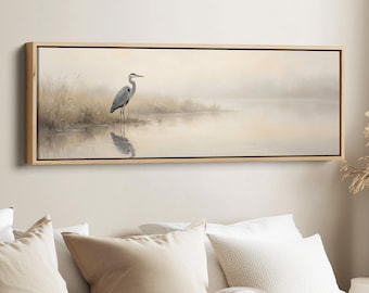 May include: A tranquil landscape painting depicting a grey heron standing in still water. The artwork is framed in natural wood, with soft, muted colours creating a peaceful atmosphere. The heron's reflection is visible in the water.