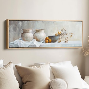 Op de afbeelding: Een ingelijst stilleven schilderij met twee crèmekleurige keramische vazen, een grijze kom en verschillende kleine sinaasappels. Een witte doek is onder de vazen gedrapeerd. Gedroogde bloemen en bladeren zijn rechts gerangschikt. Het schilderij zit in een licht houten frame.