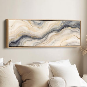 May include: Abstract art print in a rectangular wooden frame. The artwork features flowing, marbled patterns in shades of cream, gold, and gray, with subtle gold accents. The piece is a long, horizontal canvas, suitable for a bedroom or living room.