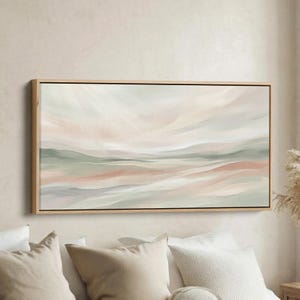 Könnte beinhalten: Abstrakte Landschaftsmalerei in einem Holzrahmen. Das Kunstwerk zeigt weiche Pinselstriche in Rosa-, Grün- und Beigetönen, die eine ruhige, hügelige Landschaft darstellen. Das Gemälde ist über weißen Kissen angebracht.