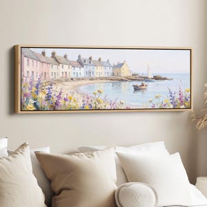 May include: A panoramic landscape painting in a light wood frame depicting a coastal village with colourful houses, a sandy beach, and a small boat on the water. The foreground features a vibrant array of purple, yellow, and white wildflowers.