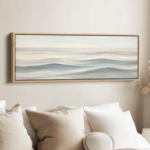 Puede incluir: Una impresión en lienzo rectangular que presenta una escena oceánica abstracta. La obra de arte representa suaves olas ondulantes en tonos azules y grises, sobre un cielo pálido. La impresión está enmarcada en un marco de madera natural, añadiendo calidez.