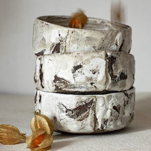May include: A stack of three white ceramic bowls with a rustic, textured finish. The bowls are stacked on a white surface with dried orange fruit scattered around them.