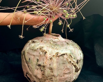 May include: A pink and green ceramic vase with a textured surface holds a dried flower with green seed pods.