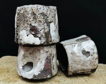May include: Three small, gray ceramic cups with white and red glaze. The cups are stacked on top of each other and are sitting on a light brown surface.