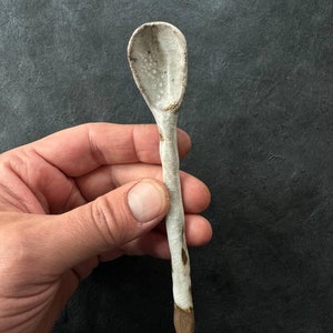 May include: A handmade ceramic spoon with a light gray bowl and handle. The handle has a textured, organic appearance with brown accents and a brown tip. The spoon is held against a dark background.